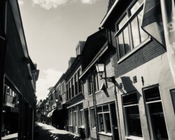 Week 18:Schaduwen A black and white, vertical photograph of a narrow cobblestone street lined with multi-story buildings. On the right side, the buildings feature several windows and a lantern mounted on the wall. A large piece of fabric or a flag hangs from an upper story on the right. Long, diagonal shadows stretch across the brick-paved ground. The top of the frame shows a sky with scattered clouds.