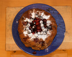Week 15: Product Fotografie An overhead view of a round, golden-brown cake on a dark blue plate. The cake is dusted with white powdered sugar and topped with a cluster of dark blue and red berries. The blue plate rests on a light-colored wooden surface. This wooden surface is placed on a larger, light-colored wooden floor.